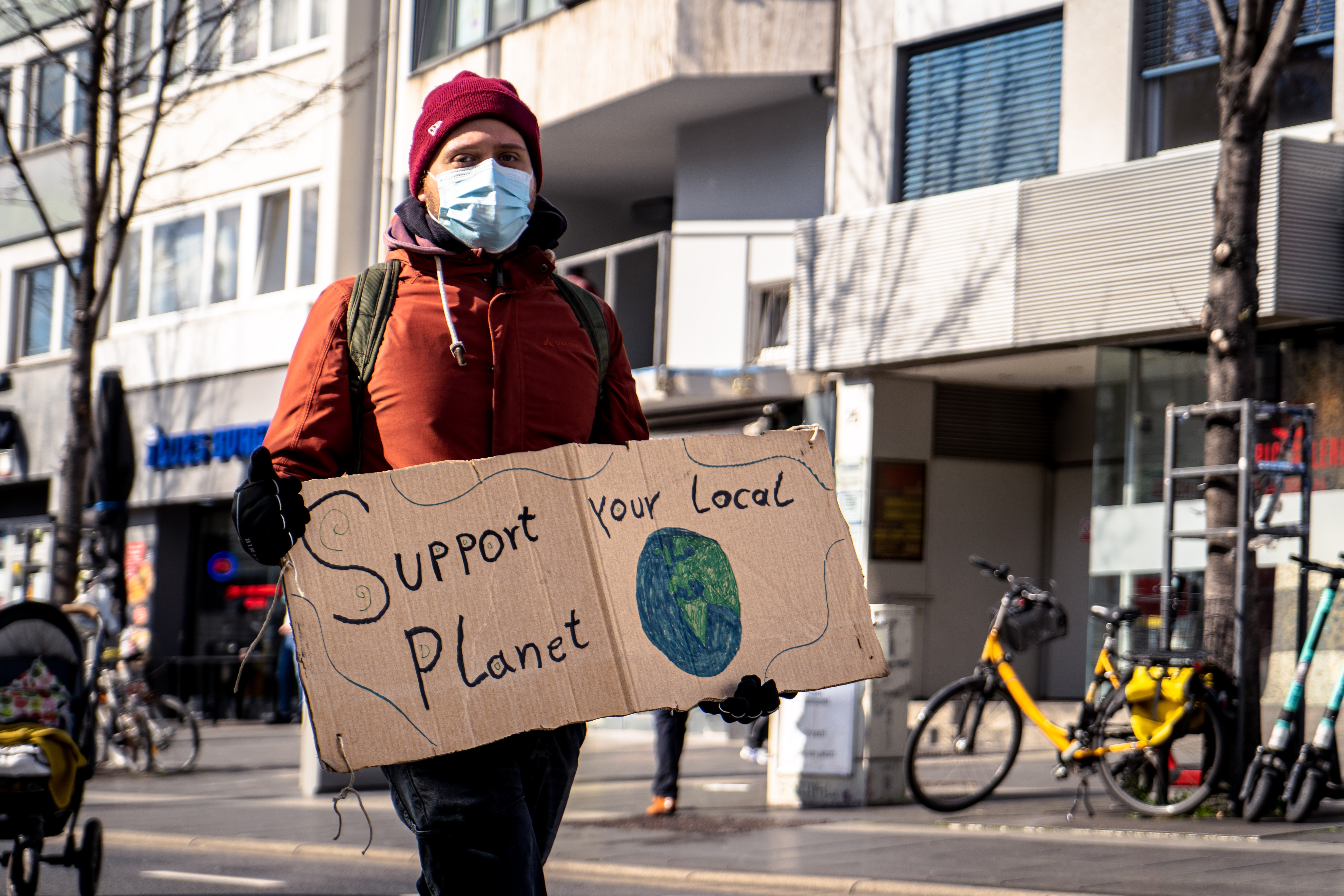 person in red, hooded winter jacket holding cardboard sign reading "support your local planet"